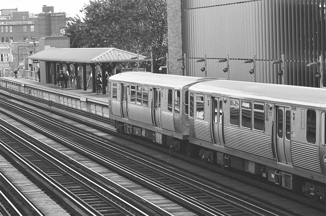 The front two cars of a six-car 5000-series Purple Line Express to the Loop set glides into Wellington station. The train is on the farthest track of a four track main line, the platform is behind the train.